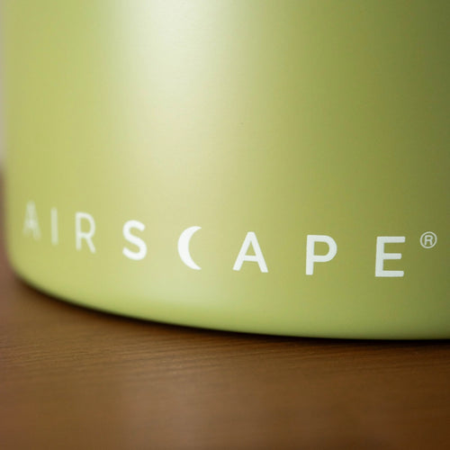 Close-up of a green container with 'AIRSCAPE' branding on a wooden surface. Airscape Matte Lichen Coffee Bean Storage Container