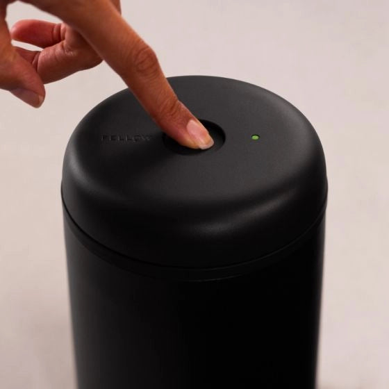 Person pressing a button on a black Fellow Atmos Electric Lid Coffee Canister with a visible brand logo.