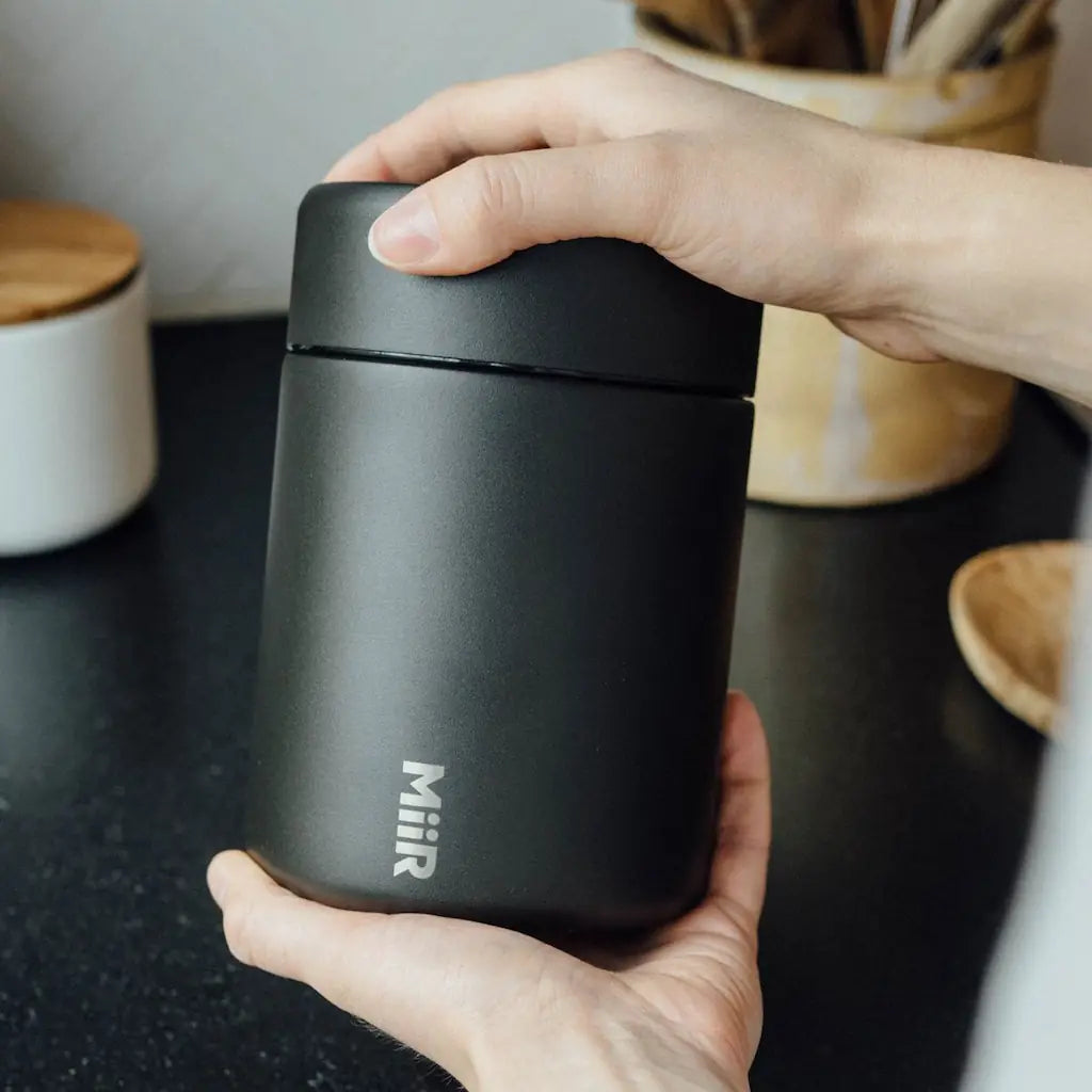 Person holding a black MiiR insulated coffee bean storage canister on a kitchen counter.