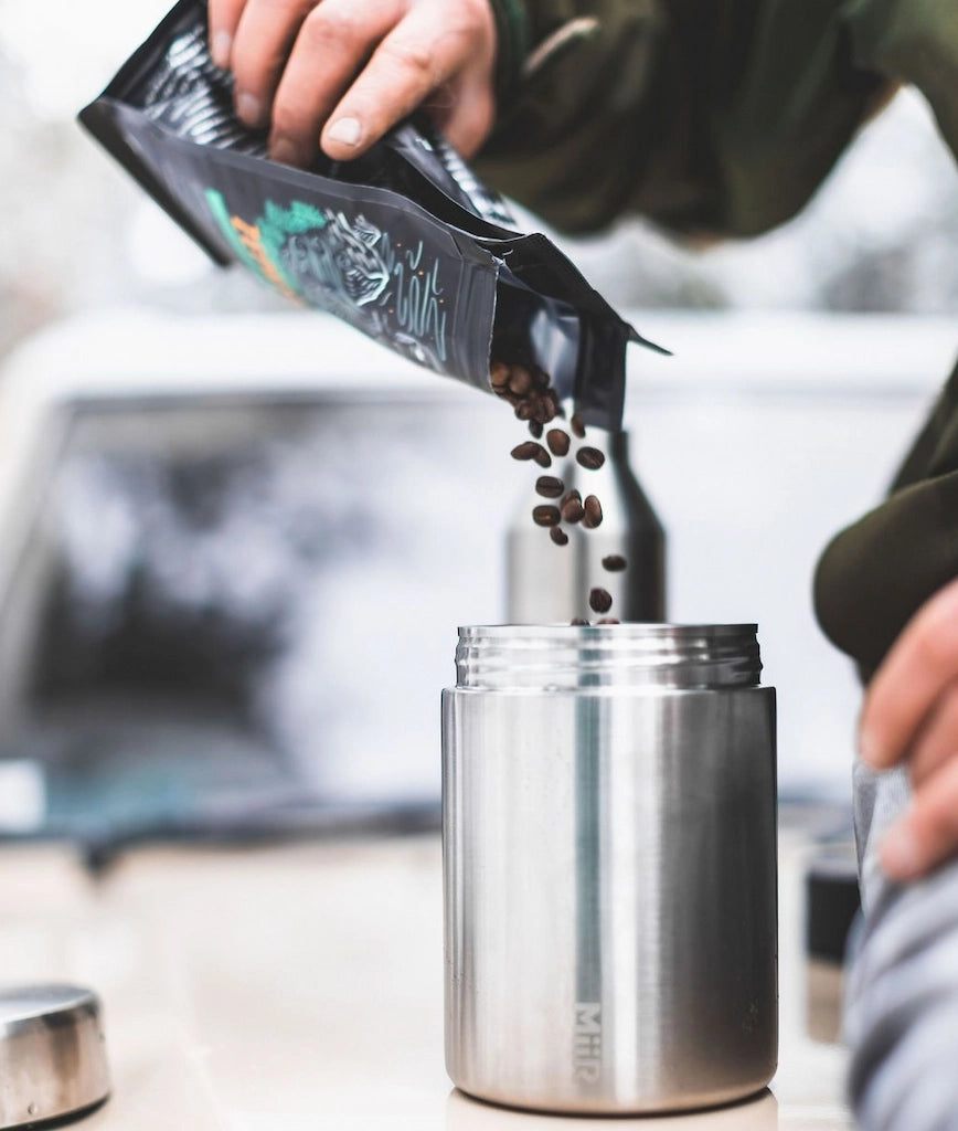 Person pouring coffee beans from a package into a silver container.